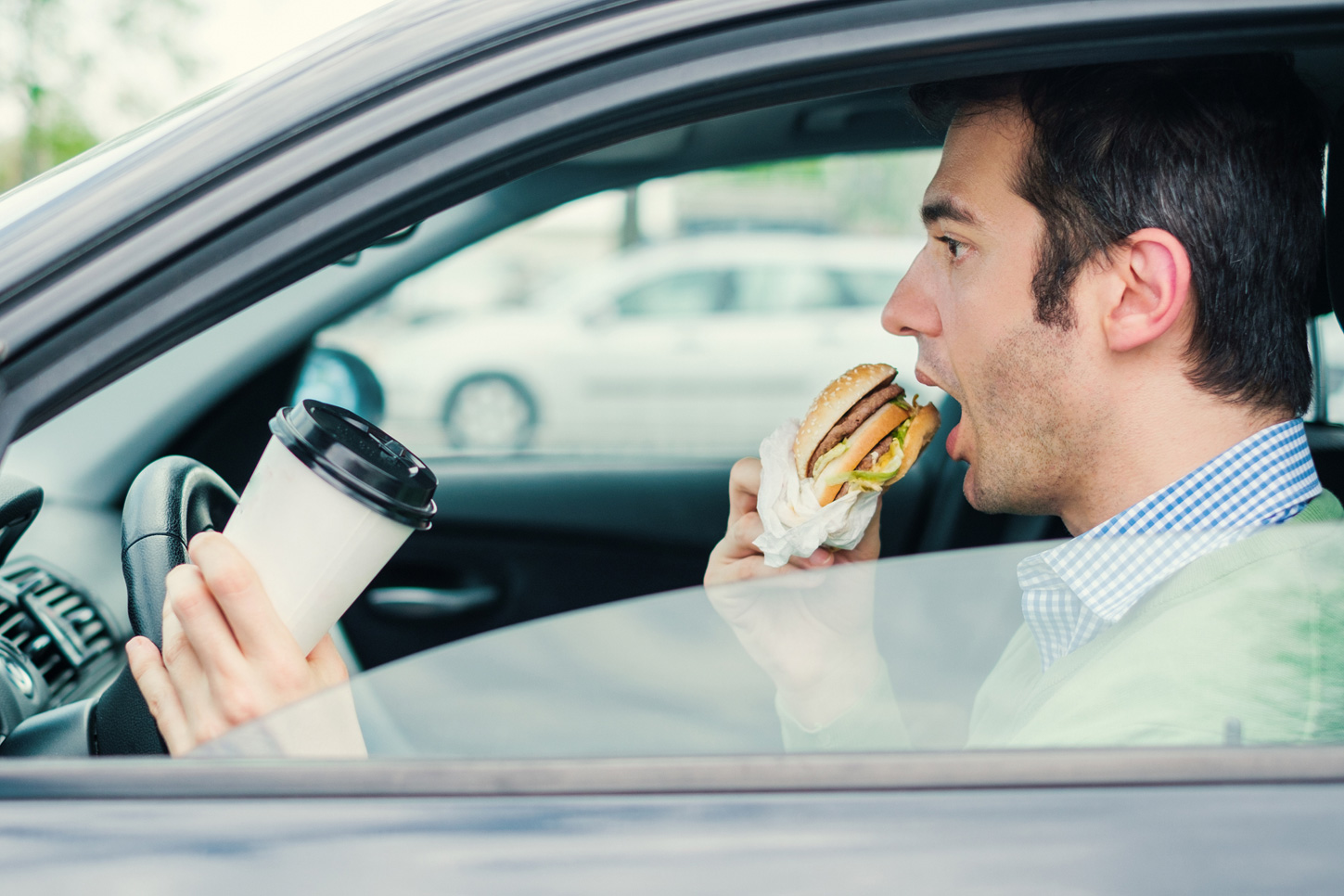 Man eating a burger and holding a coffee cup whilst trying to steer a car