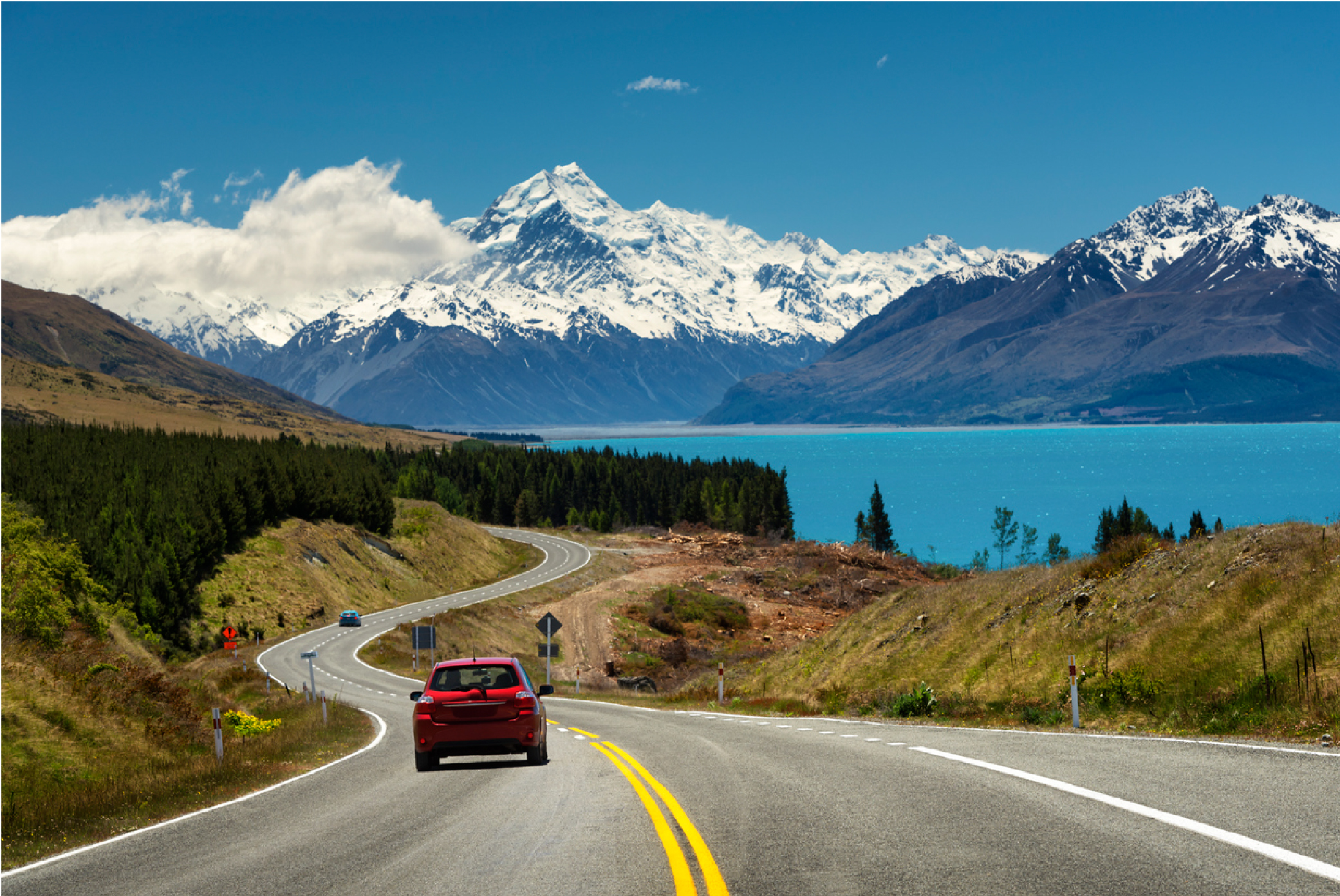 Car driving next to water and snow topped mountains